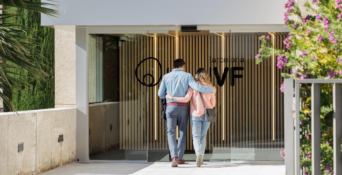 Una coppia entra insieme nell’ingresso moderno della clinica Barcelona IVF, circondato da vegetazione, pronta ad iniziare il percorso di fertilità.