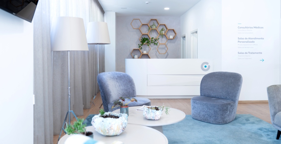 Luminoso spazio di attesa nella clinica Ferticentro a Coimbra con poltrone grigie, tavolini rotondi, tende lunghe, bancone reception bianco e decorazioni a nido d’ape sulla parete.